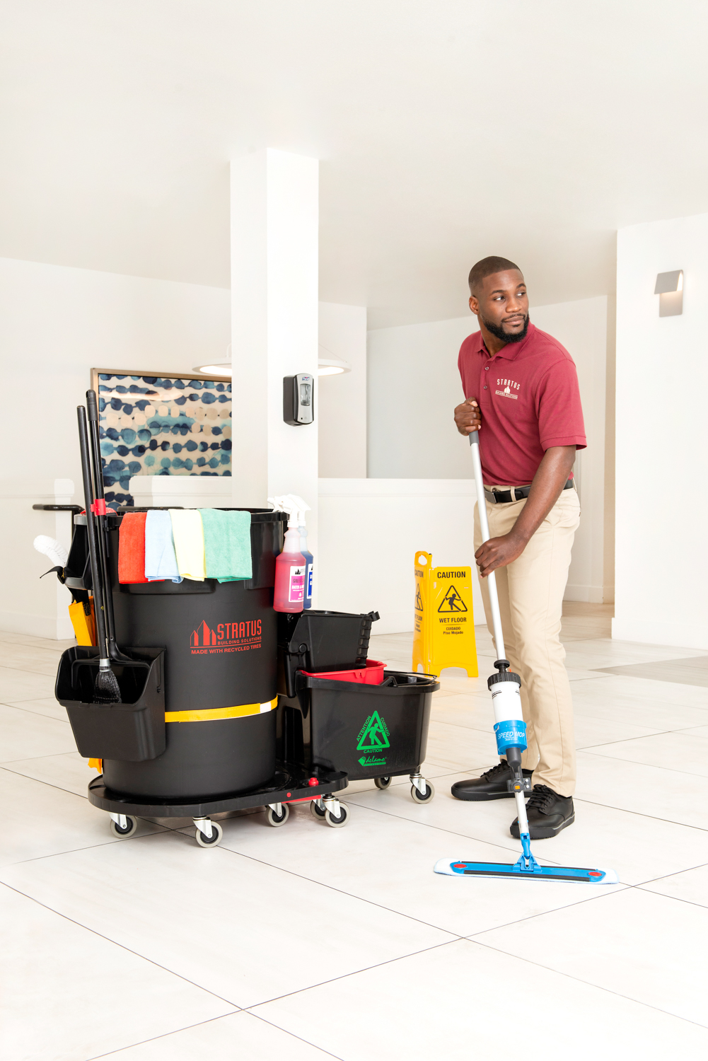 A Man in a Stratus Polo Mops a Floor with a Black Stratus Branded Trash Can and Mop Bucket Combo on Wheels Next to Him 